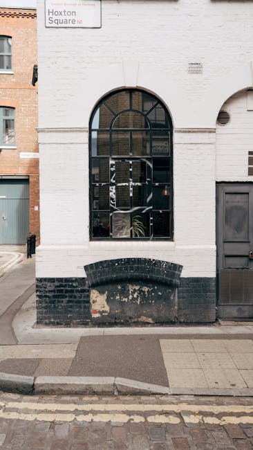 A white-painted brick building facade featuring a large, arched window with a black metal frame divided into smaller panes, revealing some indoor plants and reflections. Below the window, there is a d