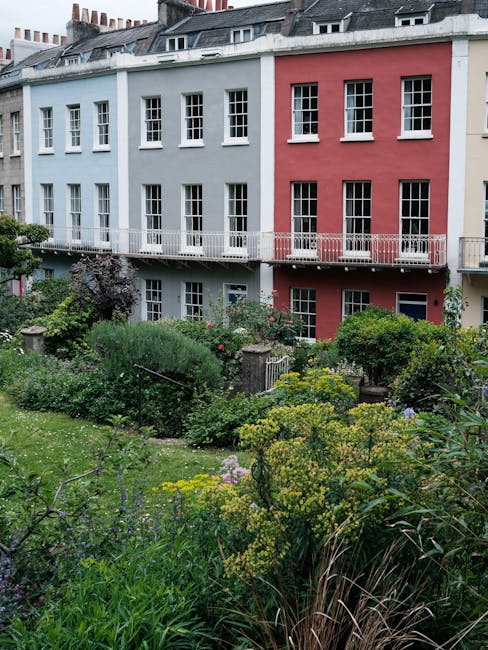 A multi-story residential building with a light blue and red facade, featuring numerous white-framed windows and a small balcony on the upper floor. In front of the building, there is a lush urban gar
